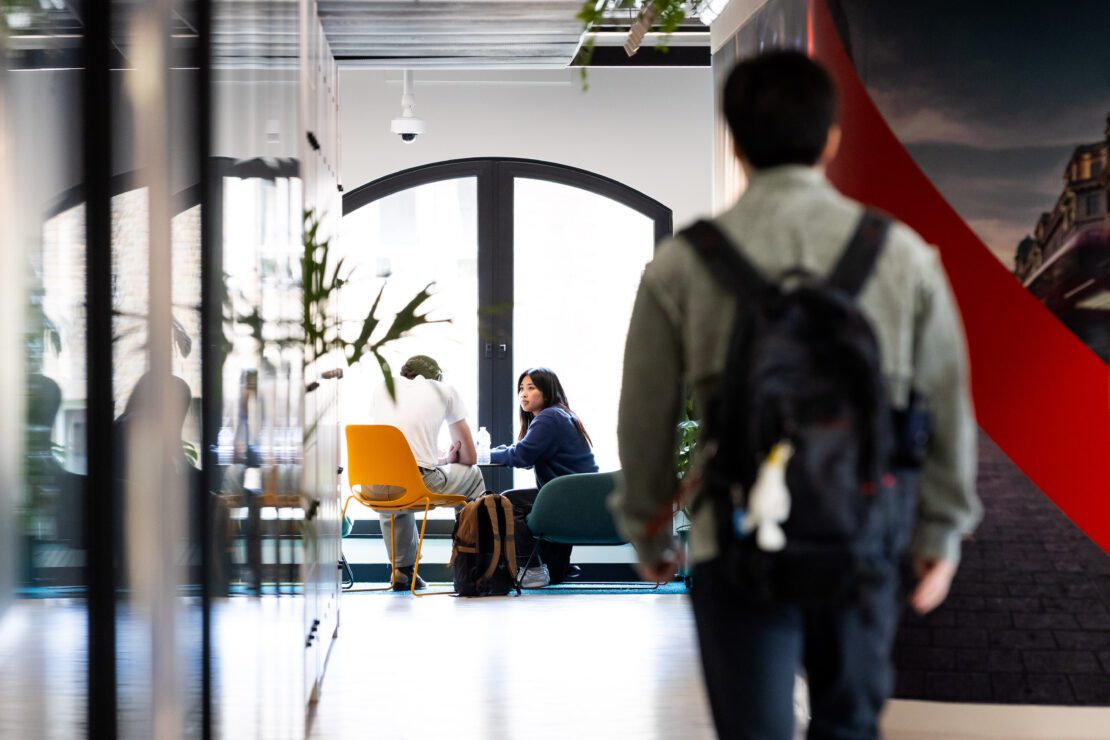 A person with a backpack walks down a hallway toward two people sitting and talking at a table near large windows in a modern office space.
