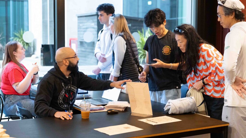 A man sits at a table with a laptop and paperwork, talking to a group of people standing in line. One woman shows something on her phone while another person writes. Others wait, and two people sit at another table in the background.