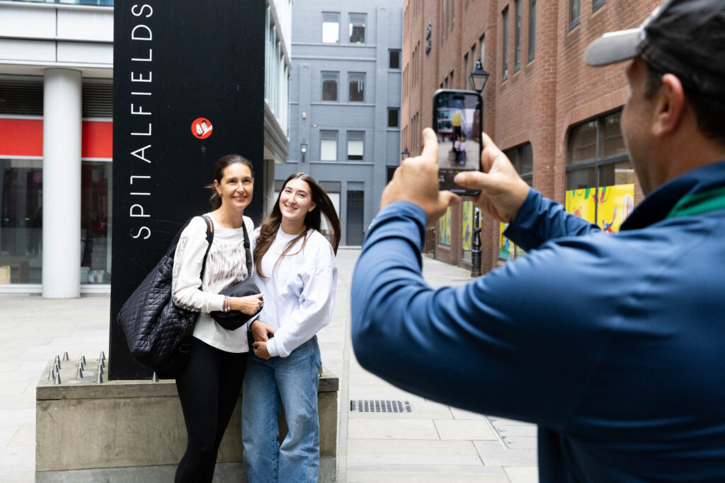 A man takes a photo with his phone of two women smiling and posing together in front of a sign that reads "Spitalfields" on a city street.
