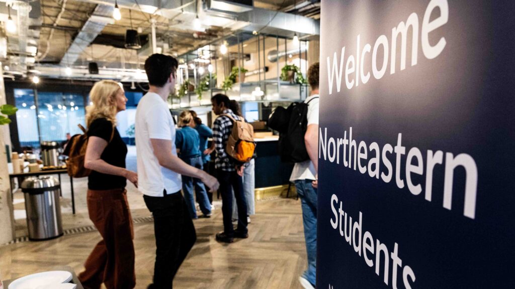 Students walk into a modern building with a large sign in the foreground that reads "Welcome Northeastern Students." The background shows a well-lit, open space with people and greenery.
