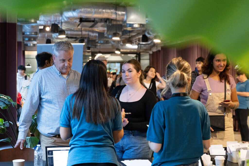 A group of people stand at a registration table, speaking with staff members in teal shirts at a busy indoor event. The background is filled with attendees and industrial-style ceiling ducts. Green leaves frame the image.