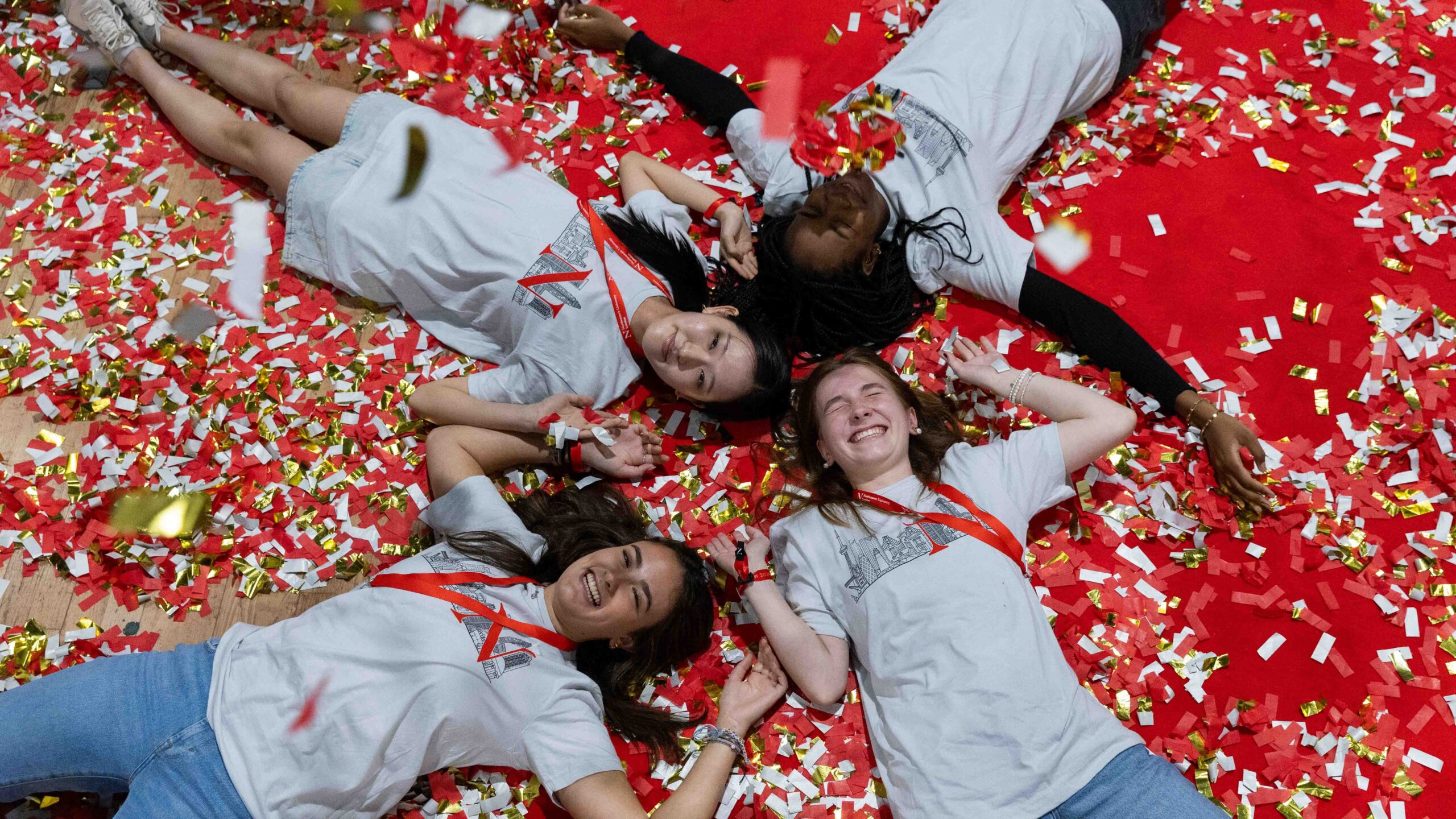Four people lie on a red floor covered in gold, red, and white confetti, smiling and wearing matching white t-shirts with red lanyards. Their heads are close together, forming a circle.