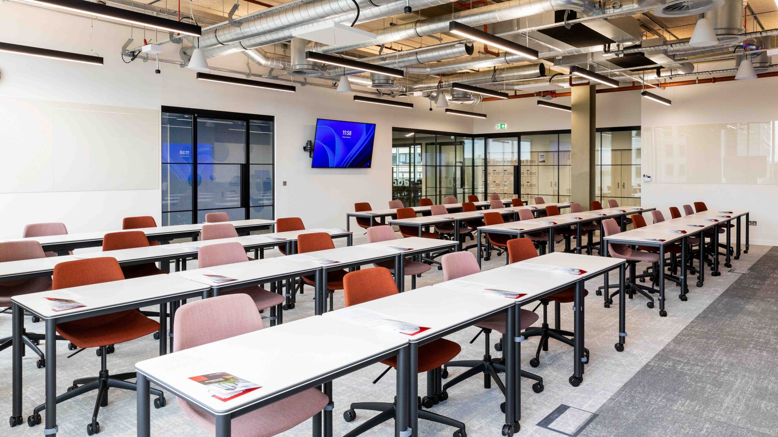 A modern classroom with rows of white tables and red chairs, each table holding a booklet. The room has large windows, a wall-mounted screen, exposed ceiling pipes, and glass walls dividing adjacent rooms.