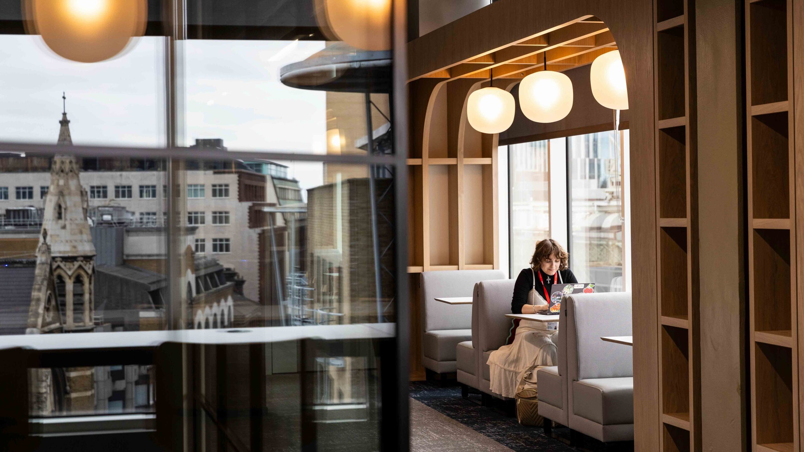 A person sits alone in a modern, well-lit booth by large windows, working on a laptop. City buildings and a church steeple are visible outside the windows. The interior features wooden accents and round hanging lights.