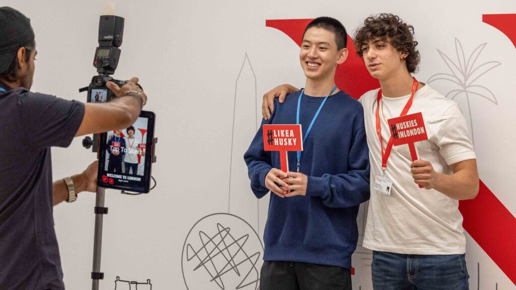 Two young men pose together holding signs while another person takes their photo. One sign reads "LIKE A HUSKY," and the other says "HUSKIES WELCOME." They stand in front of a wall with simple line art drawings.