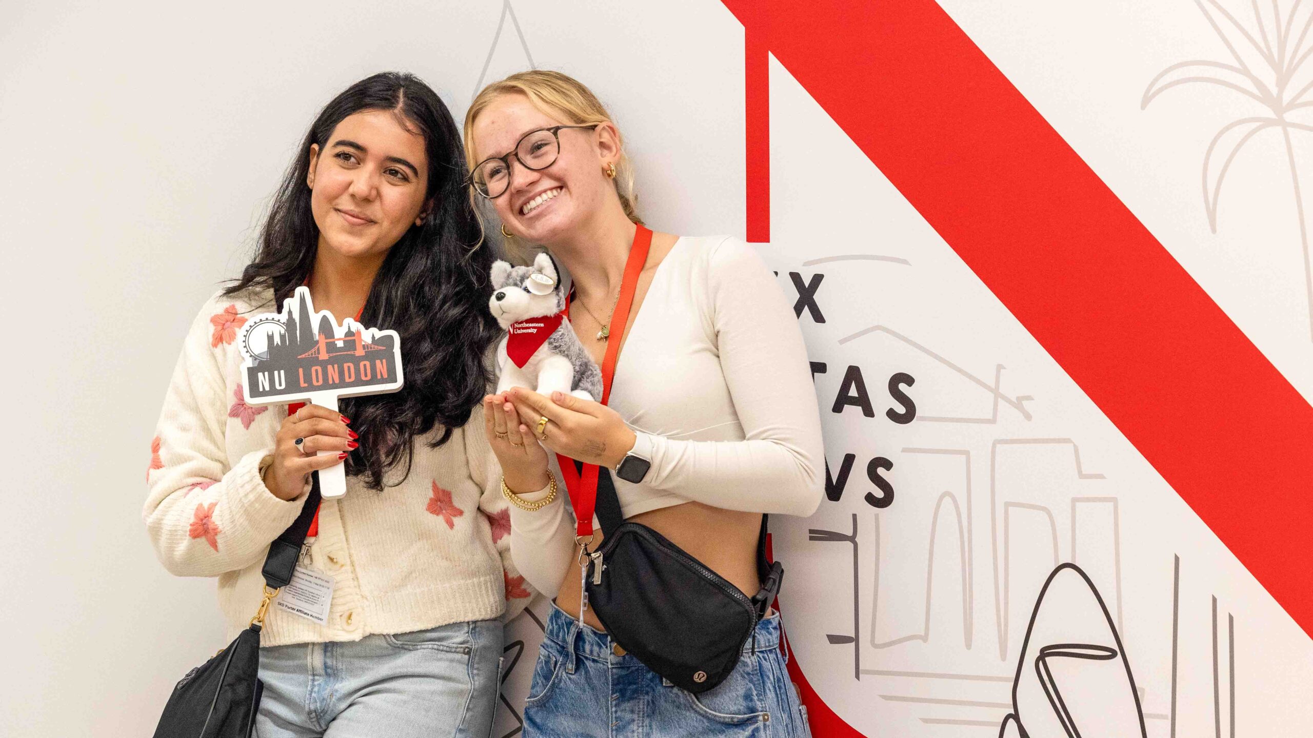 Two young women smile and pose for a photo; one holds a “NU London” sign and the other holds a stuffed husky toy. They stand in front of a white wall with red and black graphic designs.