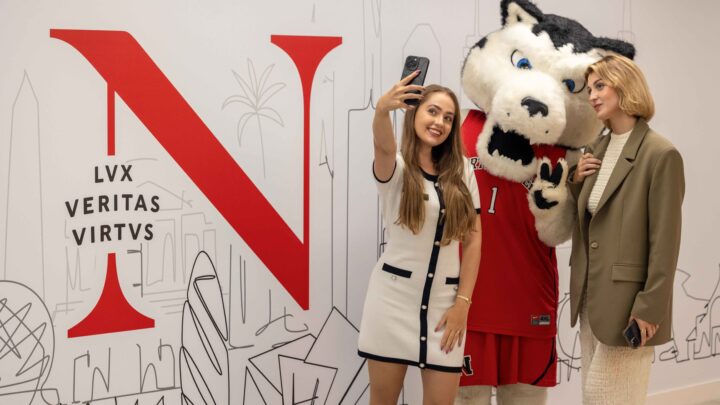 Two women pose for a selfie with a person in a husky mascot costume in front of a large red "N" and the words "LVX VERITAS VIRTUS" on a white wall with line drawings of buildings and palm trees.