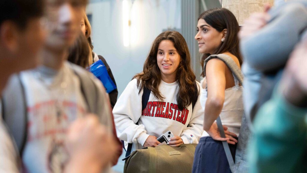 A young woman wearing a Northeastern sweatshirt stands with a group of students indoors, smiling and holding her phone and backpack. Another student stands beside her, looking in the same direction.