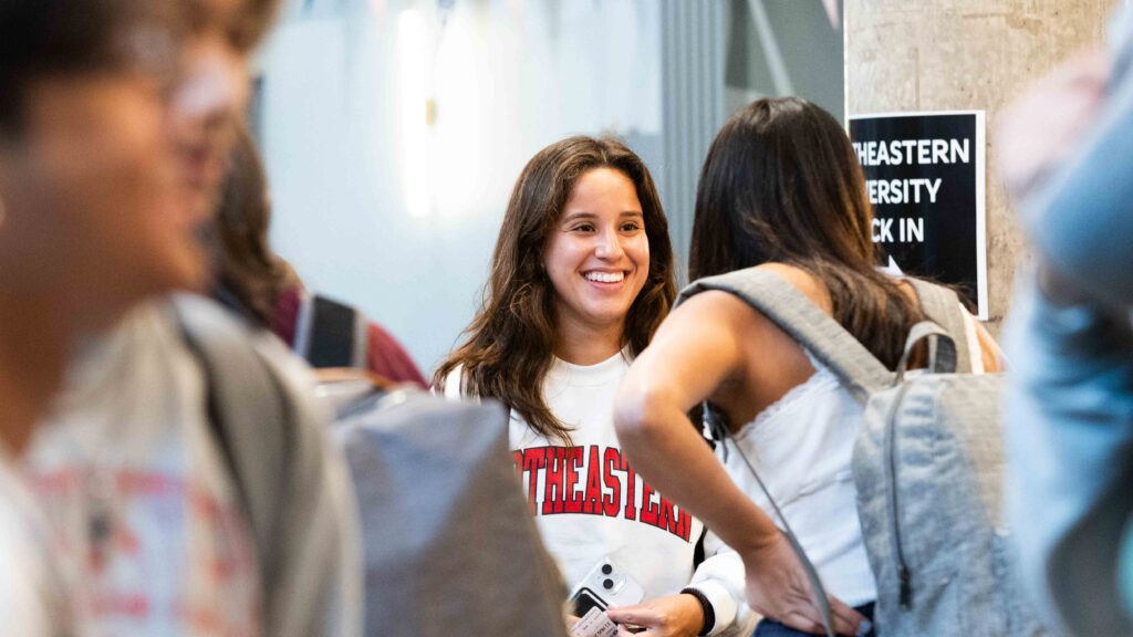 A student wearing a Northeastern sweatshirt smiles while talking to another person indoors, with a Northeastern University check-in sign visible in the background. Other students are blurred in the foreground.