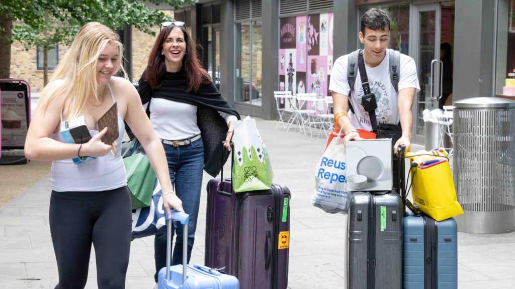 Three people smile and carry suitcases and bags along a city sidewalk, appearing to be moving in or traveling. Two women and one man are dressed casually and seem cheerful as they handle their luggage.