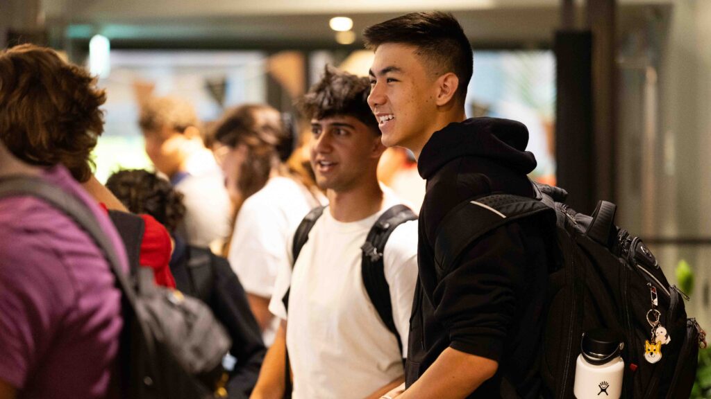 A group of young people, some wearing backpacks, stand together indoors, smiling and talking. The focus is on two people in the center, one in a white shirt and the other in a black hoodie.