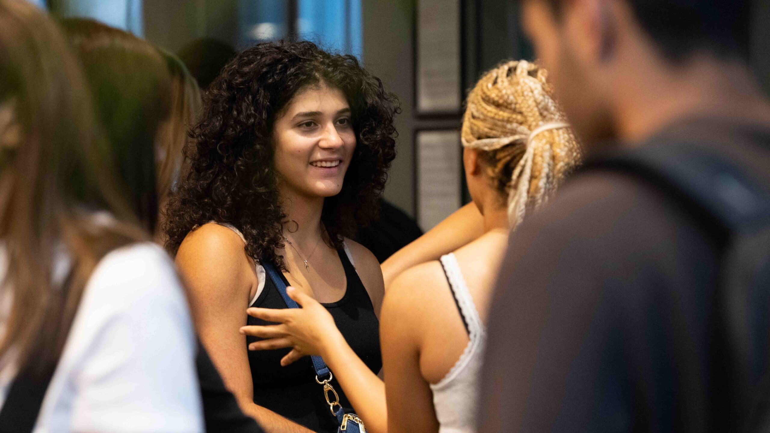 A young woman with curly hair and a black tank top smiles while talking to others in a casual indoor setting. People around her are engaged in conversation.