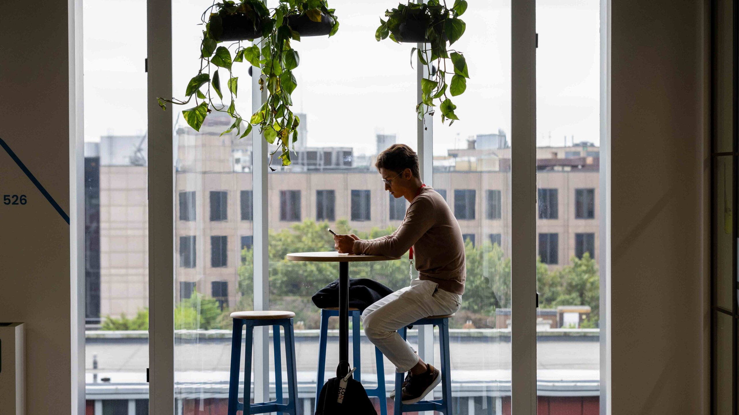 A person sits alone at a tall table by a large window, looking at their phone. Two empty stools and hanging plants are nearby, with a cityscape visible through the window.