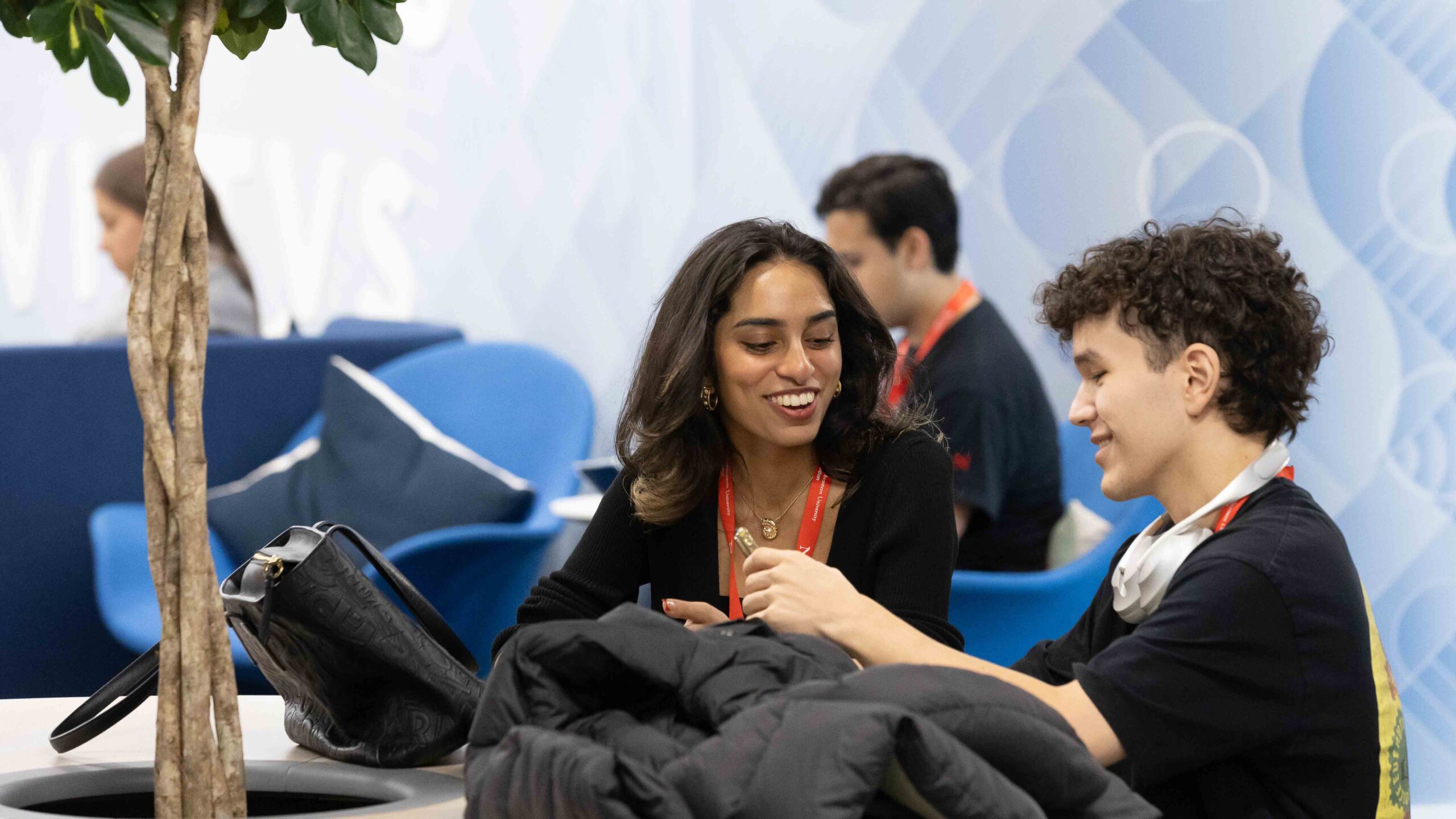 Two young people sit at a table, smiling and talking. They wear event lanyards and casual clothes. A tree is in the foreground, with modern blue furniture and other people blurred in the background.