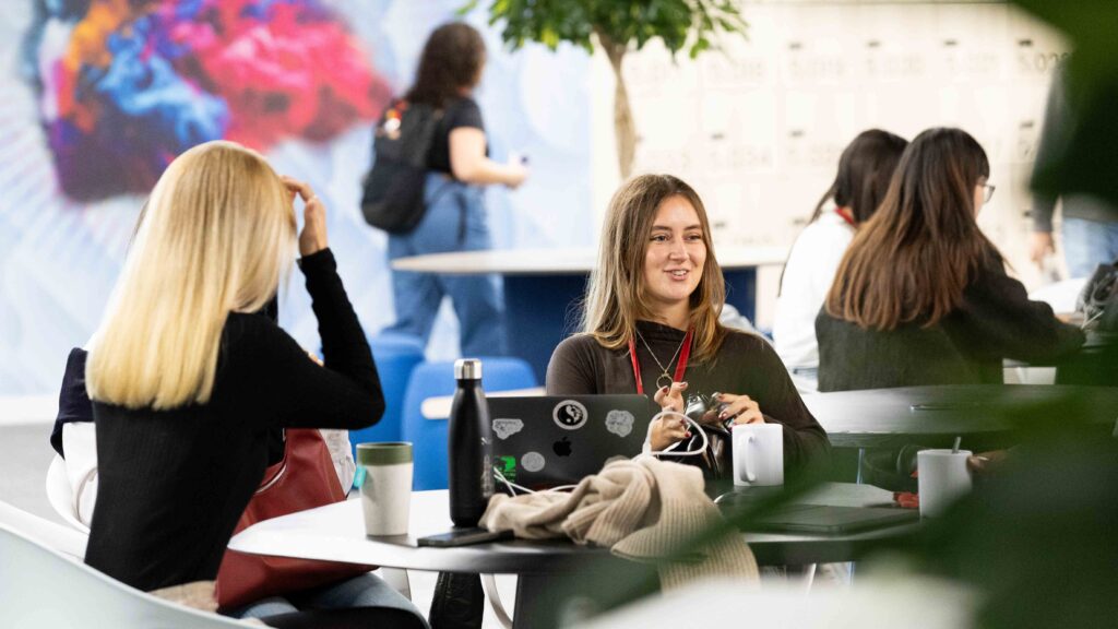 Two women sit at a round table with laptops and coffee cups, talking and smiling in a bright, modern indoor space. Other people are seated or walking in the background, with plants and colorful wall art visible.