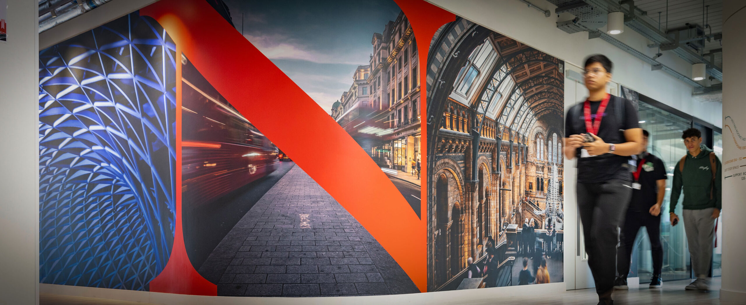 A blurred person walks past a large, colorful wall mural featuring vibrant cityscape photography and abstract geometric patterns in a modern hallway.