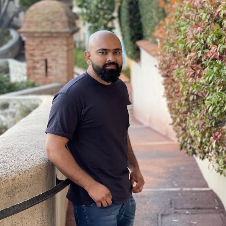 A man with a beard and shaved head, wearing a dark t-shirt and jeans, stands on a curved outdoor pathway lined with stone walls and lush greenery, looking toward the camera.