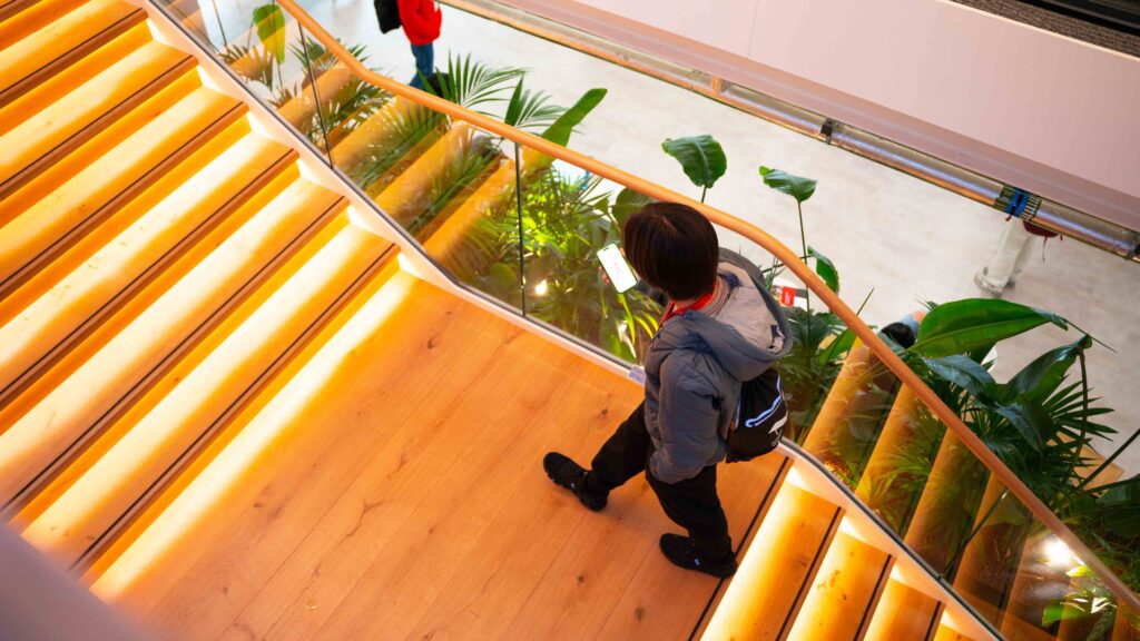 A person with short hair walks down a well-lit wooden staircase with glass railings. Green plants line the side of the stairs, and the person appears to be looking at a phone.