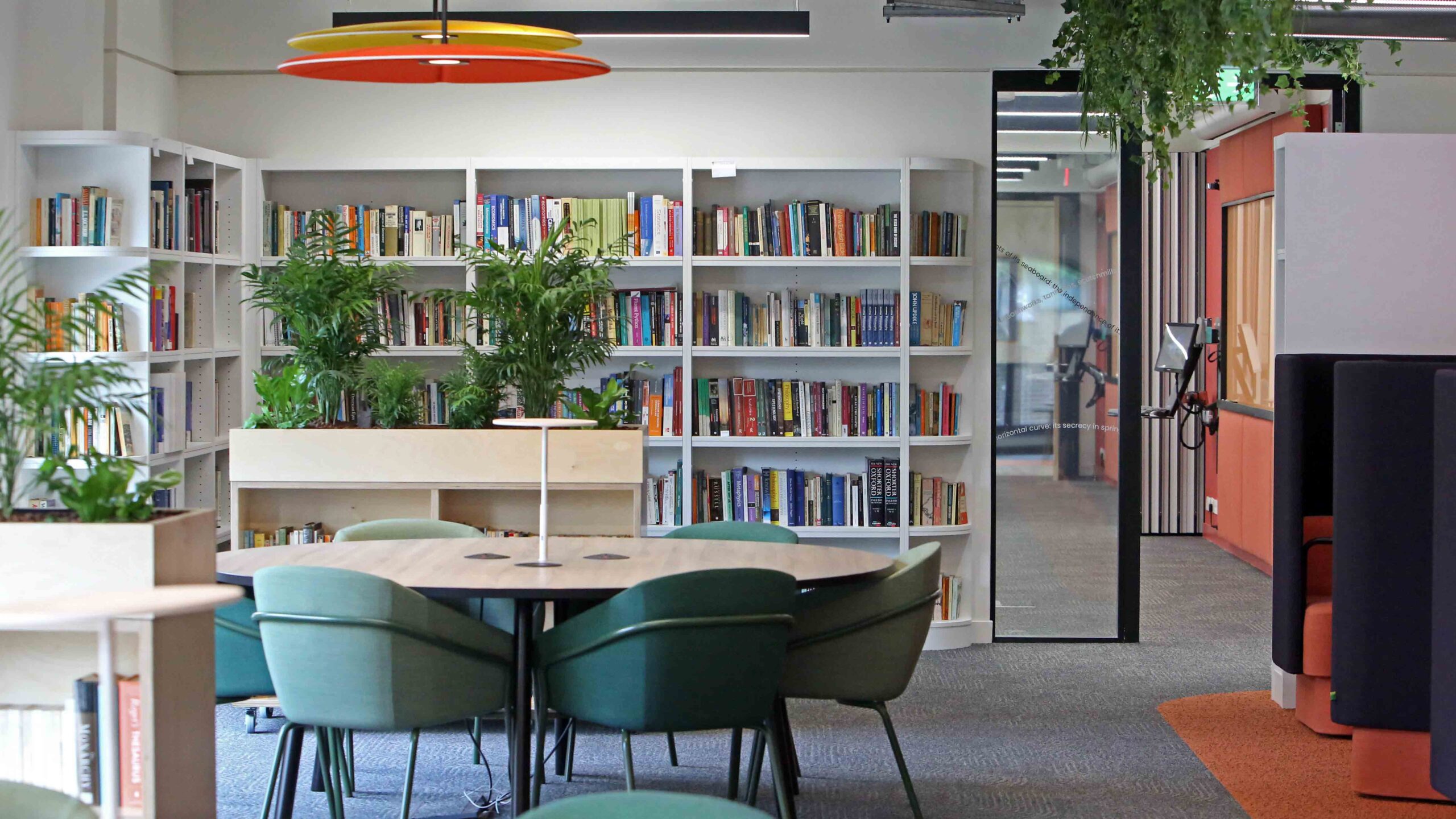 Modern library space with white bookshelves filled with books, green plants, round tables surrounded by green chairs, and large glass doors letting in natural light. The atmosphere is bright and inviting.
