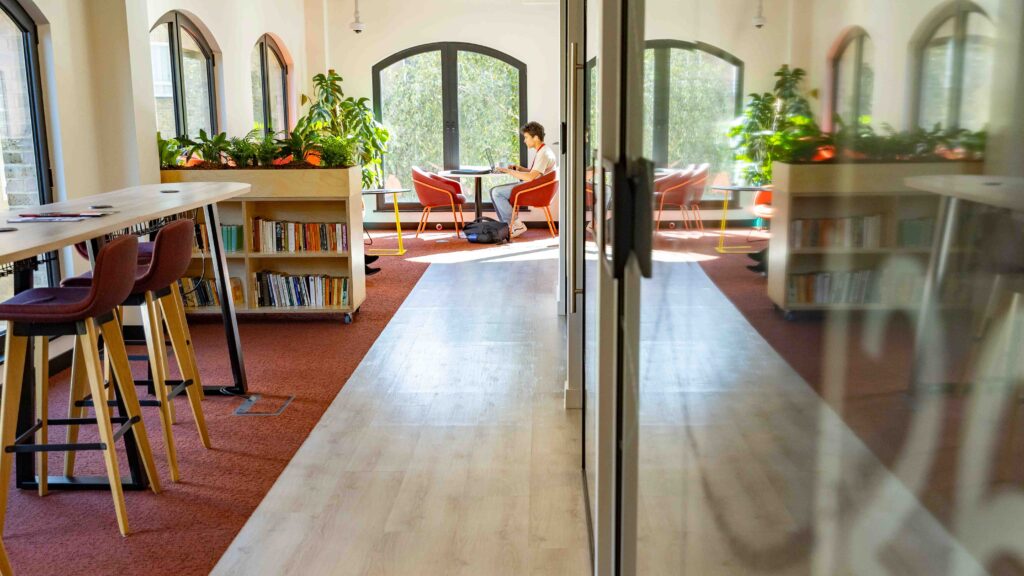 A person sits alone at a round table by large arched windows in a bright, modern room with plants, bookshelves, and high chairs along a counter. Sunlight streams in, highlighting the warm, inviting atmosphere.