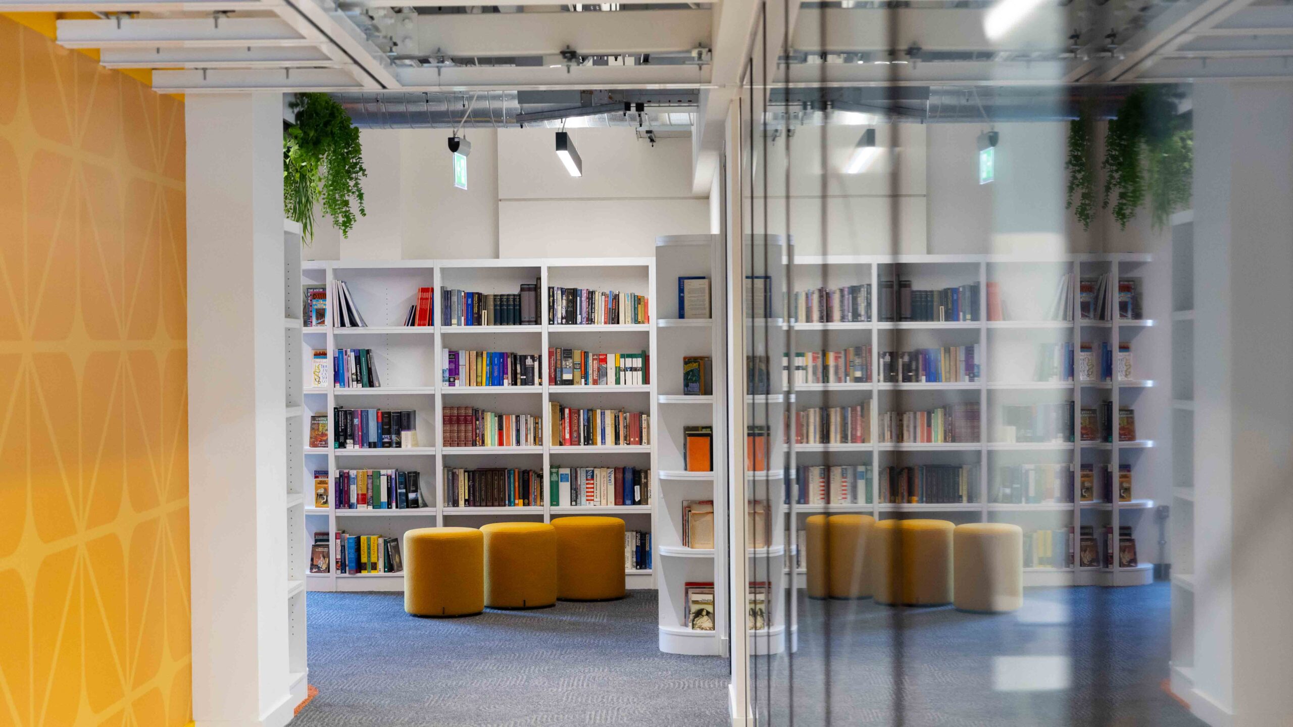 Modern library interior with white bookshelves filled with books, yellow round ottomans for seating, and reflected glass walls. The space is bright with overhead lighting and some green plants hanging from the ceiling.