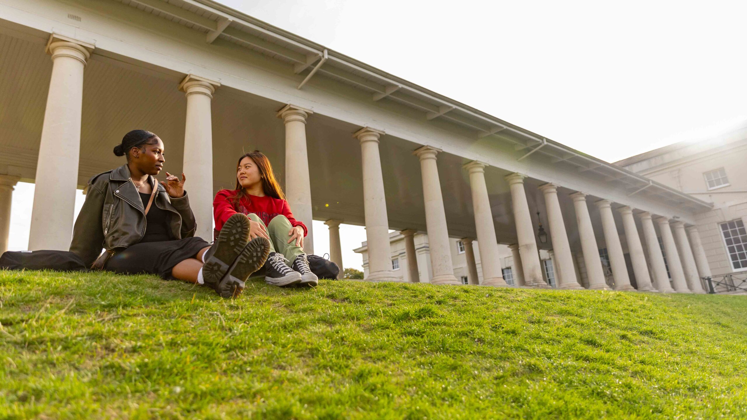 Two young women sit on a grassy hill, talking and relaxing in front of a building with tall white columns on a sunny day.