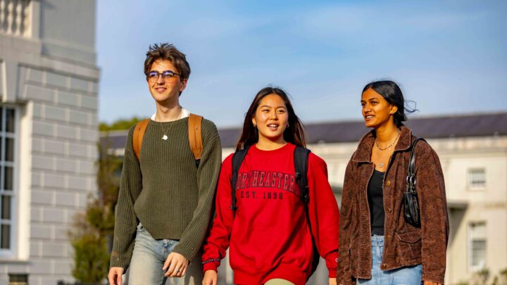 Three students walk together outdoors, smiling. One wears a green sweater and jeans, another wears a red Northeastern University sweatshirt, and the third wears a brown jacket. Buildings and blue sky are visible in the background.