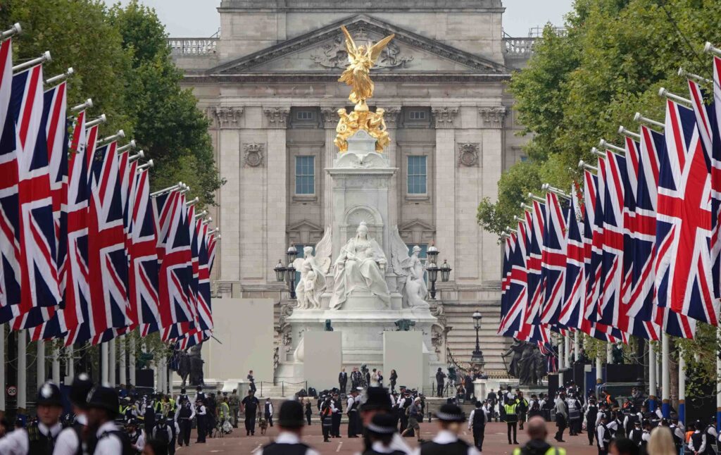 Rows of Union Jack flags line a wide street leading to the Victoria Memorial and Buckingham Palace, with people and police officers gathered along the road. Trees frame the scene on both sides.