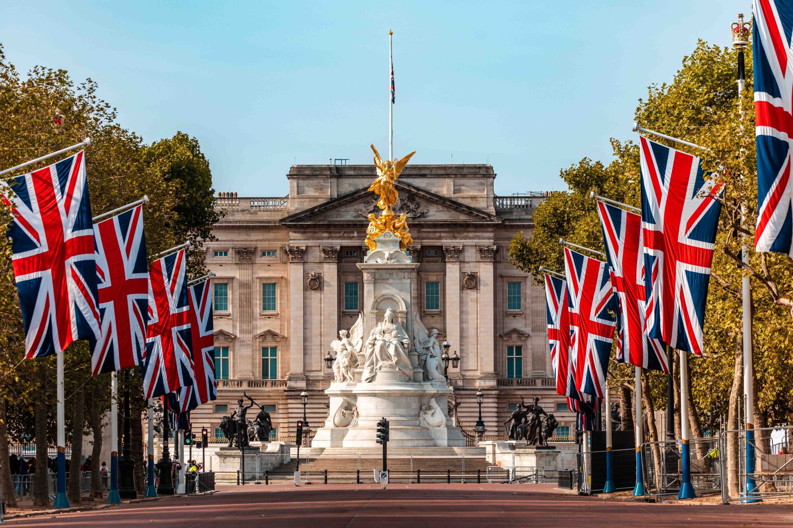 Buckingham Palace with the Victoria Memorial in front, lined by trees and large Union Jack flags on both sides of the road, under a clear sky.