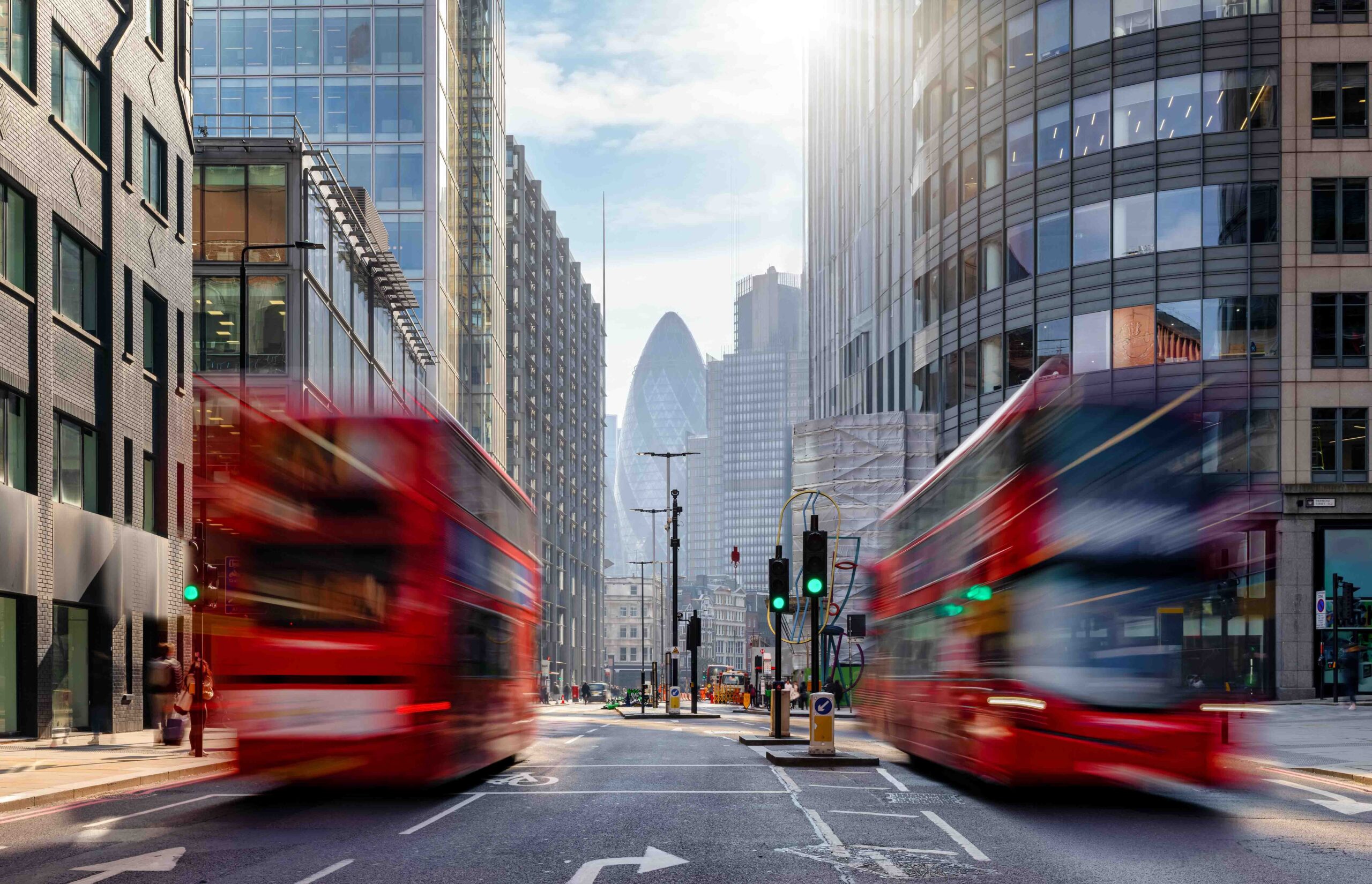 A city street in London with two red double-decker buses in motion, surrounded by tall modern buildings and the Gherkin skyscraper visible in the background under a bright sky.