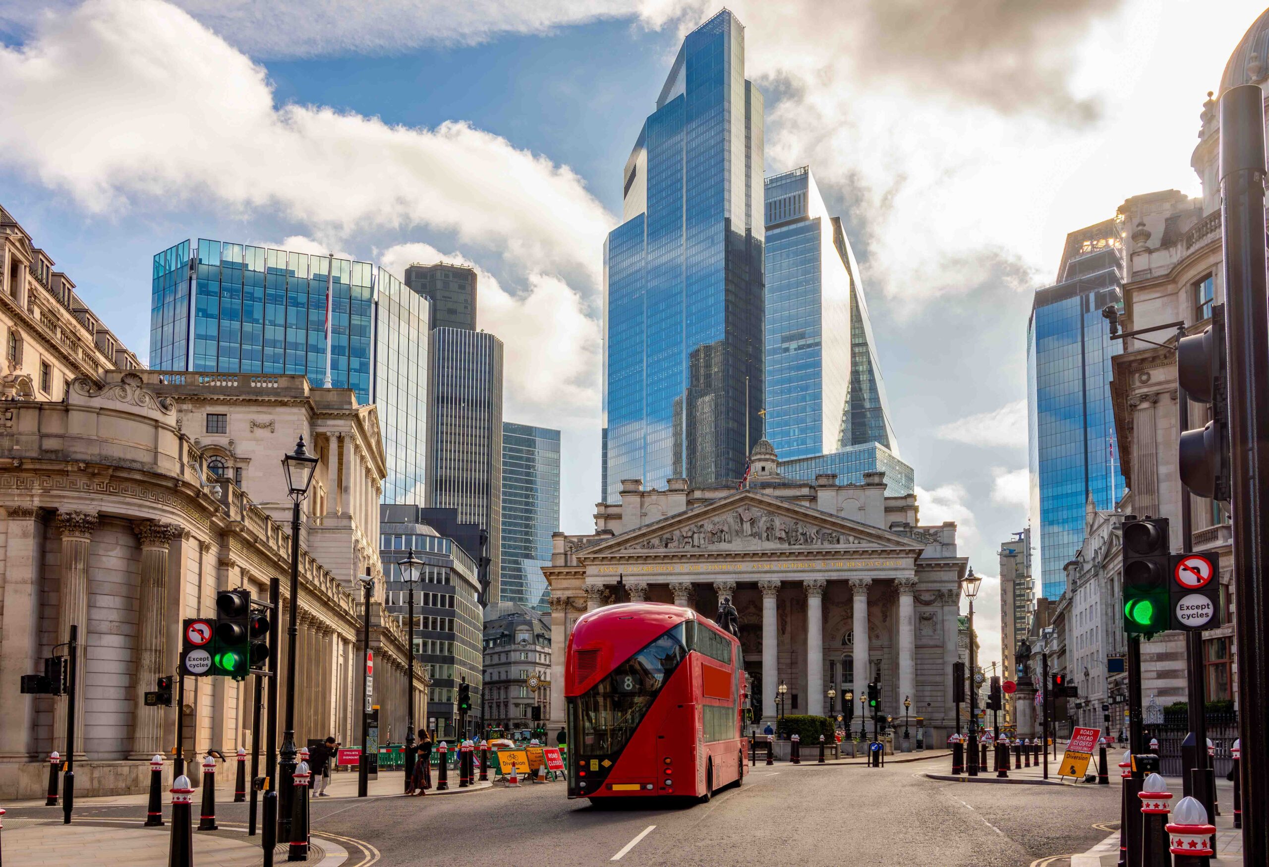 A red double-decker bus drives in front of the historic Royal Exchange building, surrounded by modern skyscrapers in London’s financial district.