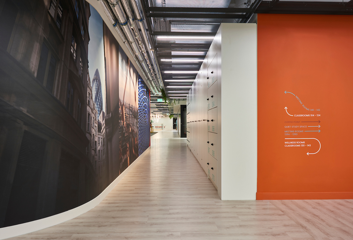 Modern hallway with light wood floors, a large mural of a cityscape on the left wall, white lockers on the right, and a bright orange wall with white directional signage further down the corridor.