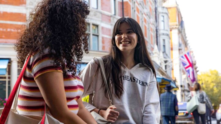 Two young women stand on a city street, talking and smiling. One wears a striped shirt, the other a light hoodie. Buildings and a British flag are visible in the background.