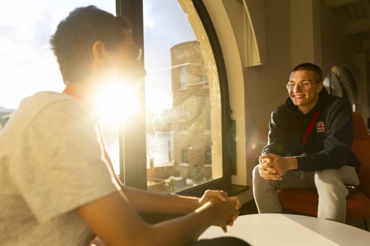 Two people sit facing each other by a large window with sunlight streaming in. One person is smiling, wearing glasses and a hoodie; the other is in partial silhouette due to the bright light.