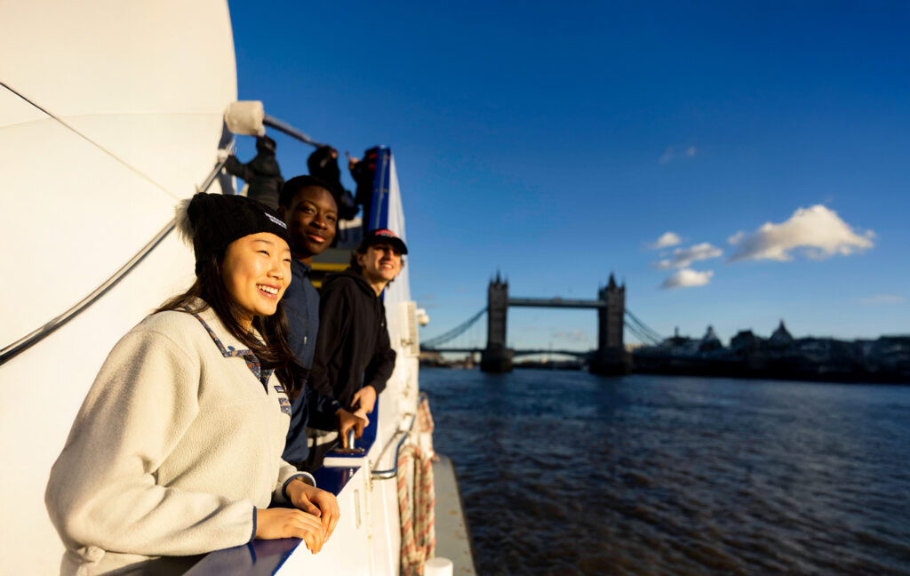 Three young people smile and look out from a boat on the River Thames, with the Tower Bridge visible in the background under a bright, clear blue sky.