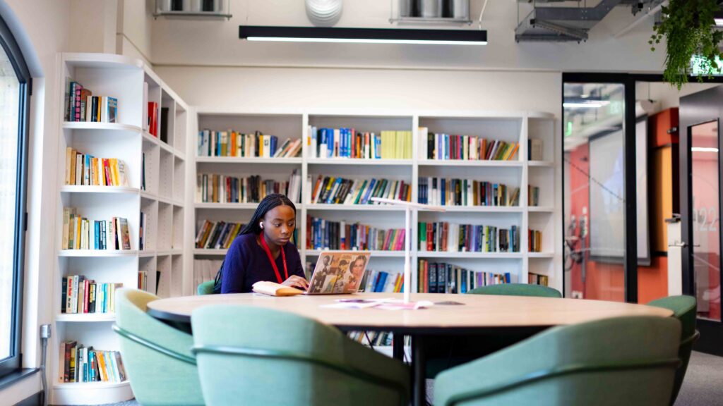 A woman sits alone at a round table in a modern library, reading a book. She is surrounded by bookshelves filled with books, and the space is bright with contemporary decor.