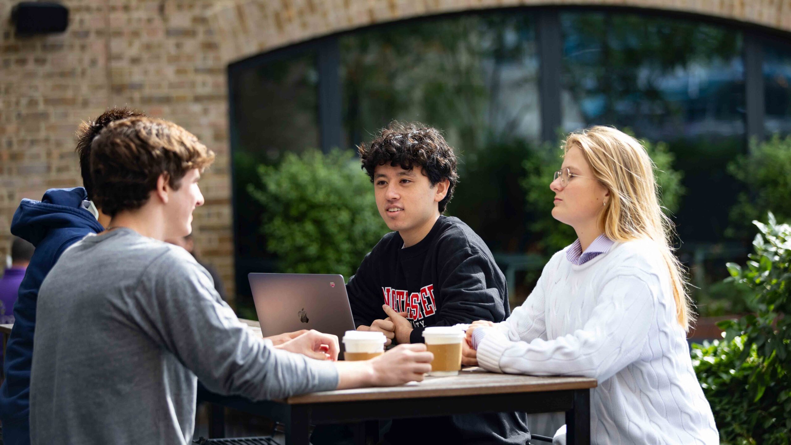 Three young adults sit at an outdoor table with coffee cups, having a conversation. One person has a laptop open. They are surrounded by greenery and a brick building is in the background.