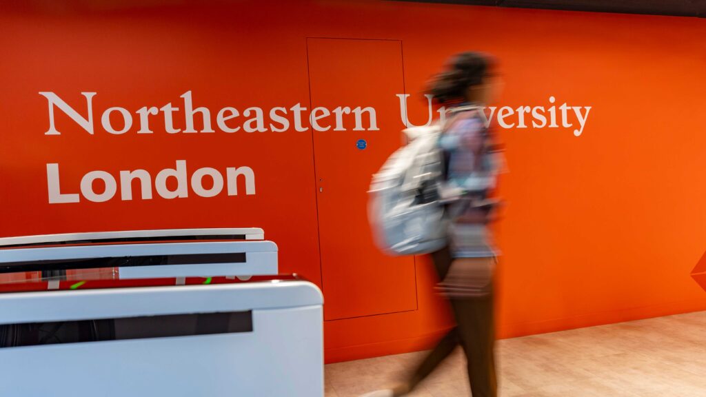 A person with a backpack walks past a bright orange wall with "Northeastern University London" written on it in white letters.