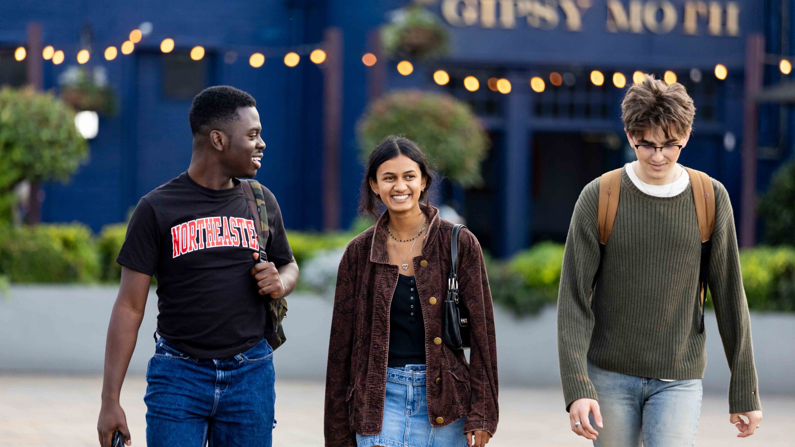 Three young adults walk outdoors, smiling and talking. One wears a "NORTHEASTERN" t-shirt, another has a brown jacket, and the third wears a green sweater. String lights and greenery are visible in the background.