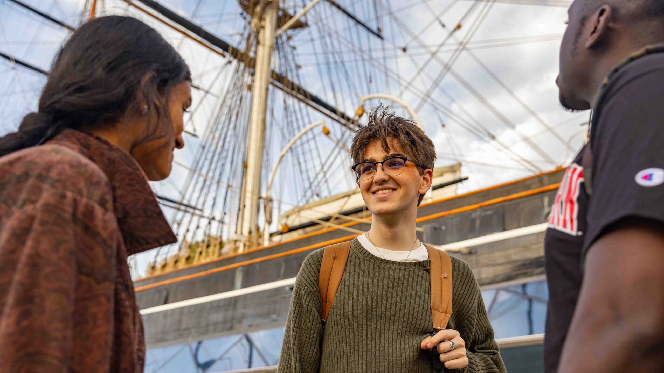 Three people stand outdoors talking and smiling near a historic sailing ship with tall masts and rigging visible in the background. One person wears glasses and a backpack.