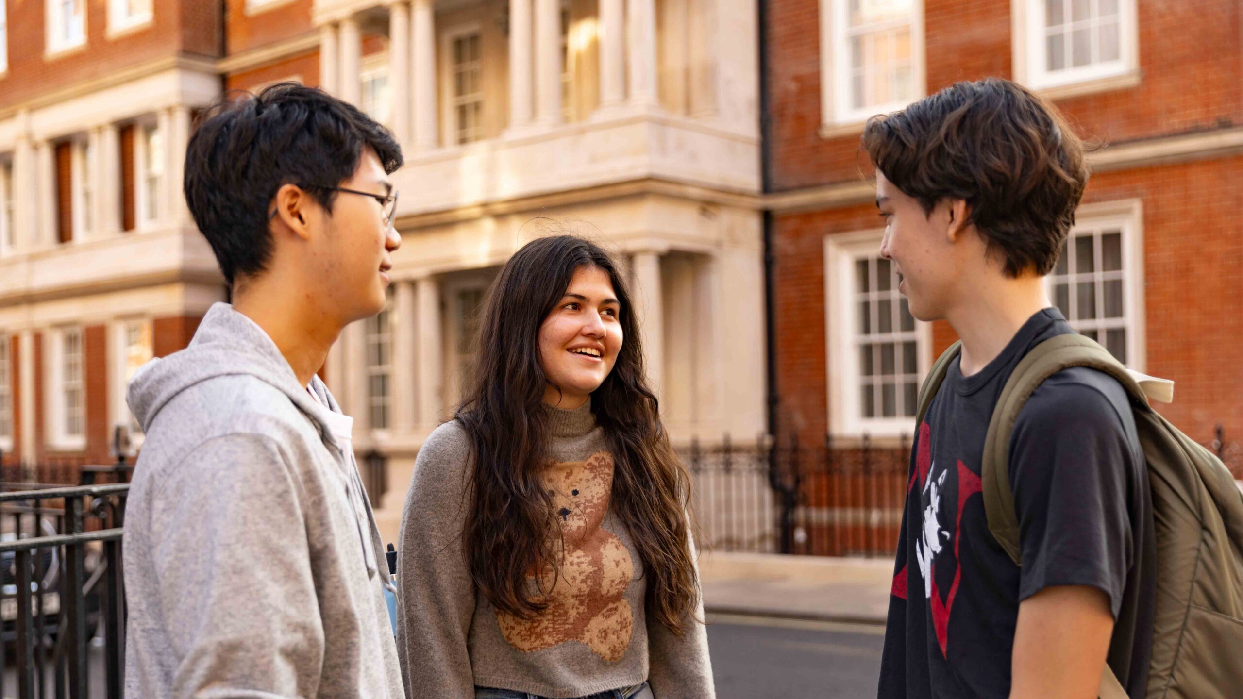 Three young people stand outside a brick building, talking and smiling. One person has a backpack, and they all appear to be enjoying their conversation on a sunny day.