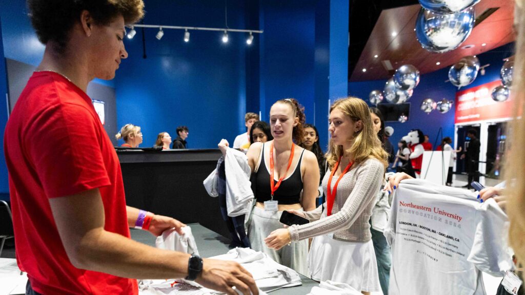A group of students wearing red lanyards receive Northeastern University T-shirts from a staff member at an indoor event, surrounded by others and shiny silver decorations.