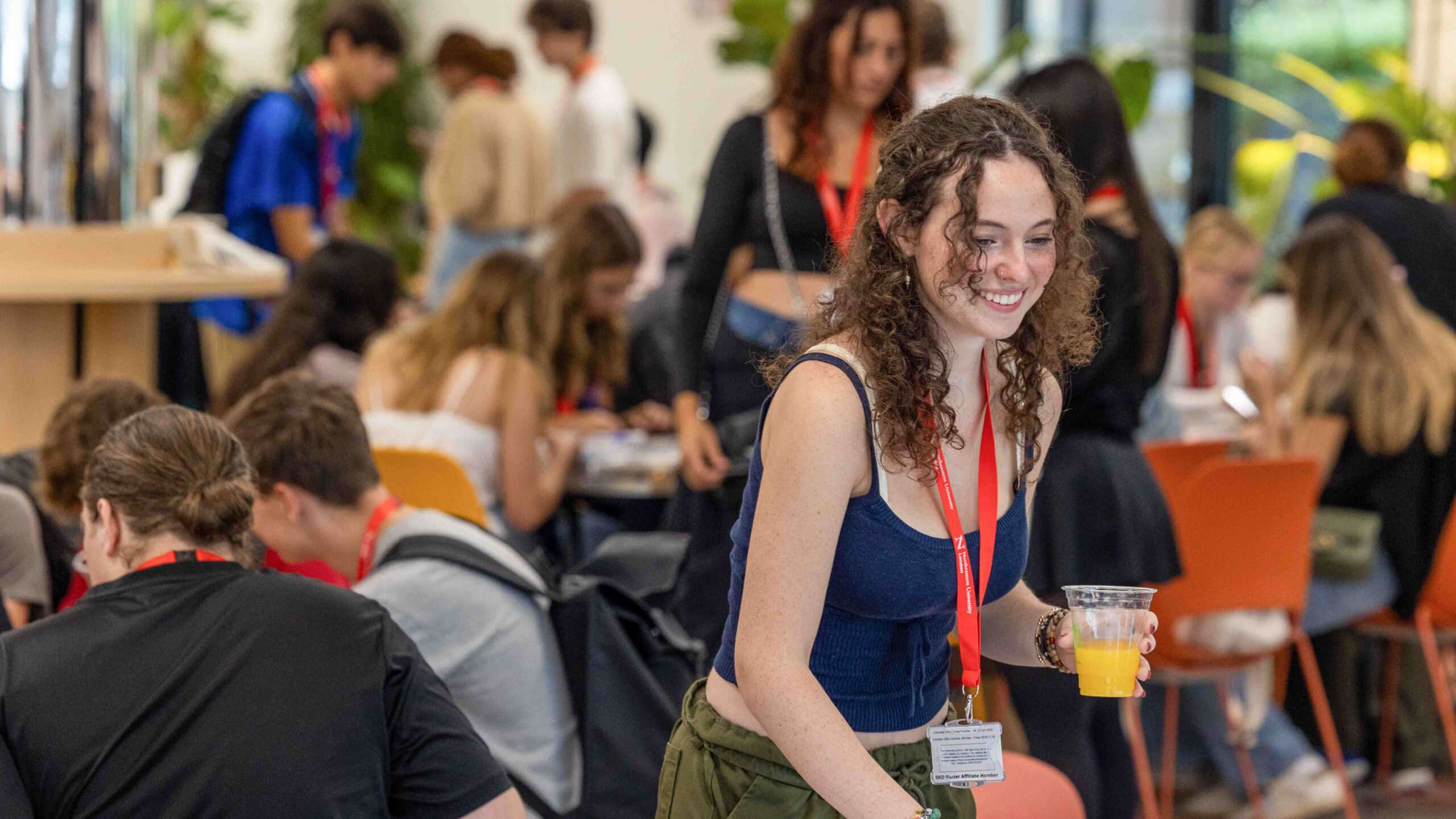 A young woman with curly hair and a red lanyard smiles while holding a cup of orange juice in a busy, brightly lit room filled with people sitting and talking.