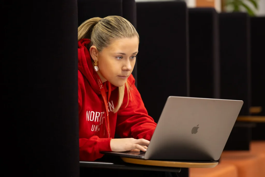 A young person wearing a red hoodie and lanyard sits at a desk, focused on using a silver laptop in a quiet, modern study area, researching how to apply through Clearing.
