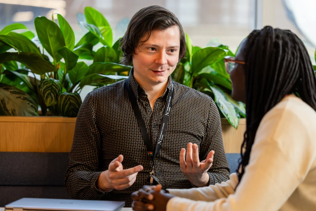Two people are sitting indoors, facing each other and having a conversation about how to apply through Clearing. One person gestures with their hands while talking, and there are green plants in the background.