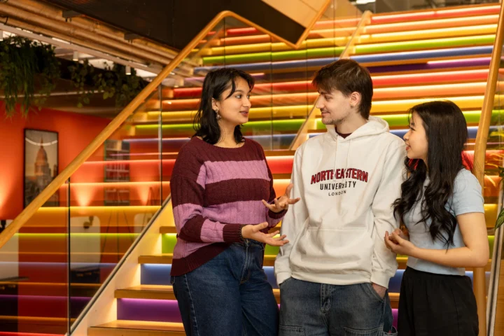 Three young adults stand and talk in front of a brightly colored, illuminated staircase, animatedly discussing Clearing FAQs. One person gestures while speaking; all appear engaged and friendly amid modern decor and indoor plants.