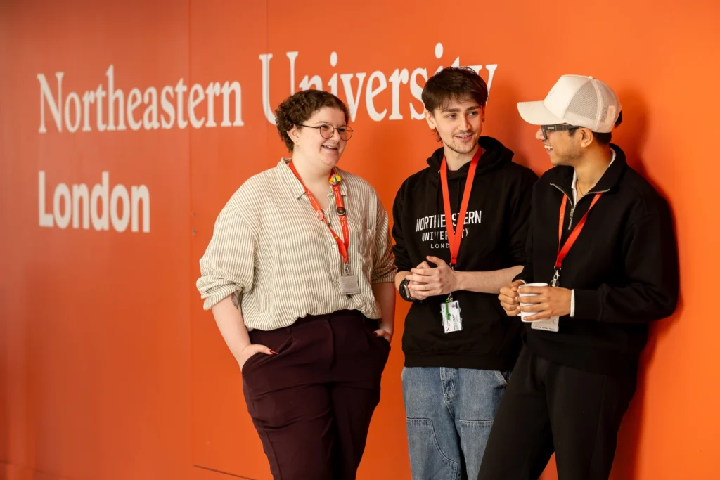 Three students with lanyards chat in front of an orange wall reading "Northeastern University London." Two are smiling while one holds a coffee cup—discussing how to apply through Clearing.