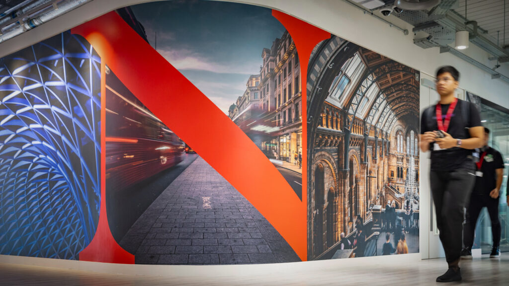 A colorful wall mural featuring city architecture and the Netflix "N" logo, with two people walking past it in a modern, well-lit building interior.