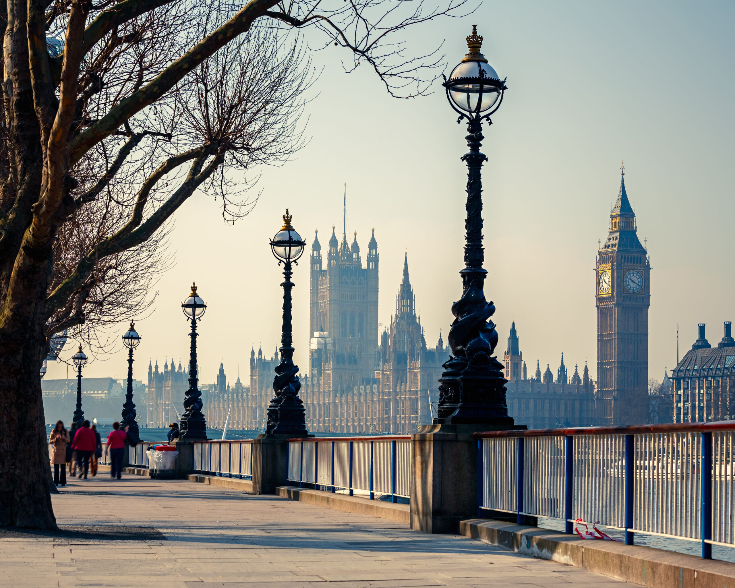 A riverside walkway in London with ornate street lamps, leafless trees, and people walking. The Houses of Parliament and Big Ben are visible in the background across the River Thames.