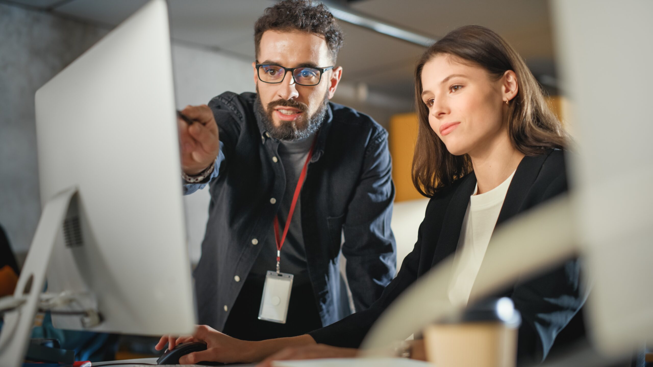 Two colleagues in an office work together at a computer. One person, standing, points at the monitor while the other, seated, uses the mouse, both focused on the screen.